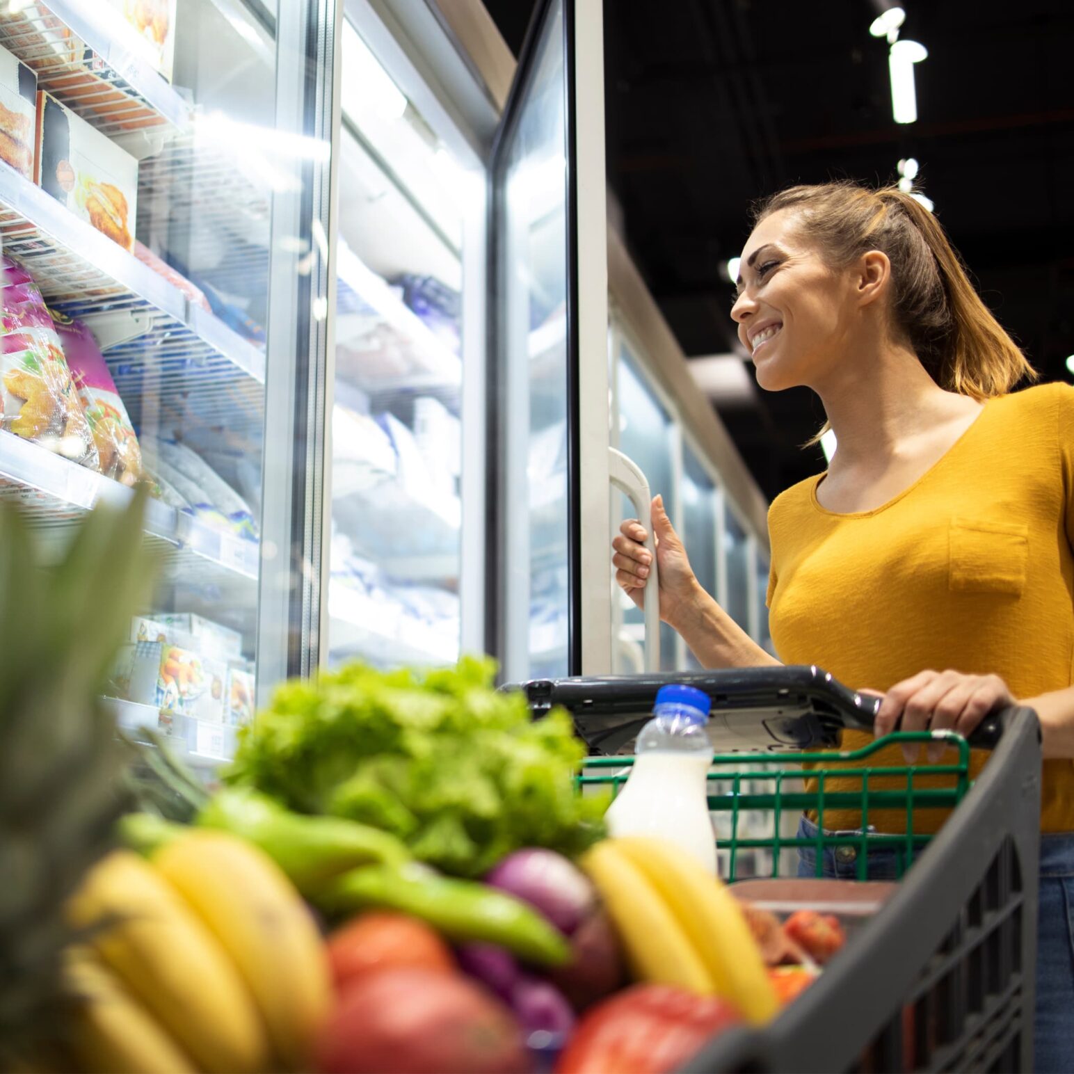 Woman holding open a grocery store freezer door with a grocery cart in front of her