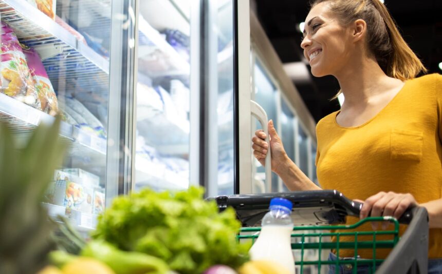 Woman holding open a grocery store freezer door with a grocery cart in front of her