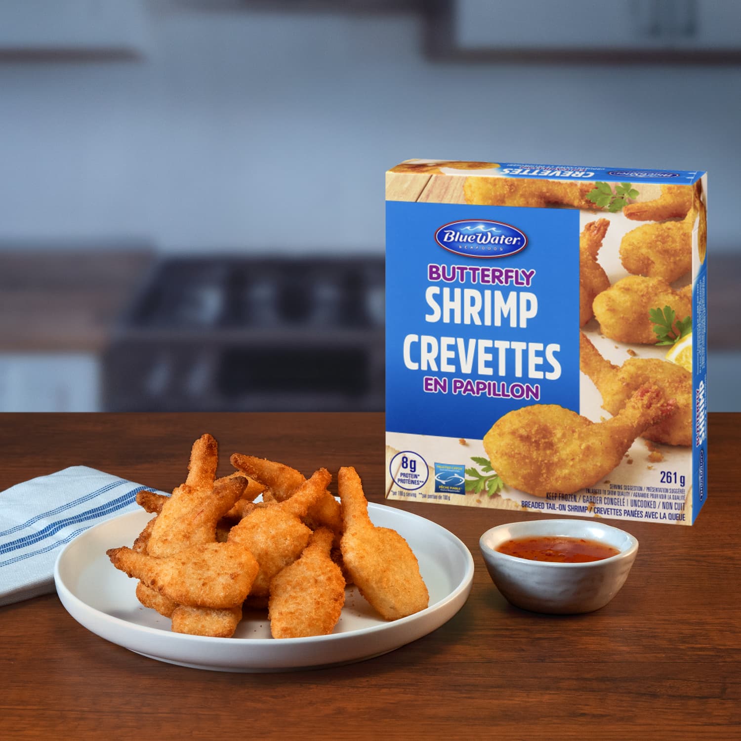 BlueWater Seafoods Butterfly shrimp on a plate next to dipping sauce and box of shrimp in a kitchen